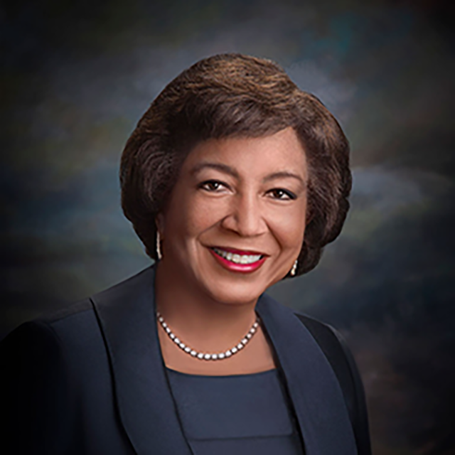 An image of a brown skinned woman with brown hair (Dr. Edith Mitchell) smiling at the camera in front of a dark multicolored background.