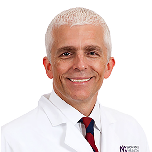 A portrait of a white haired, light skin man (Dr. Williams) smiling at the camera in front of a white background. Dr. Williams is wearing a white collar shirt, striped tie and a white lab coat.